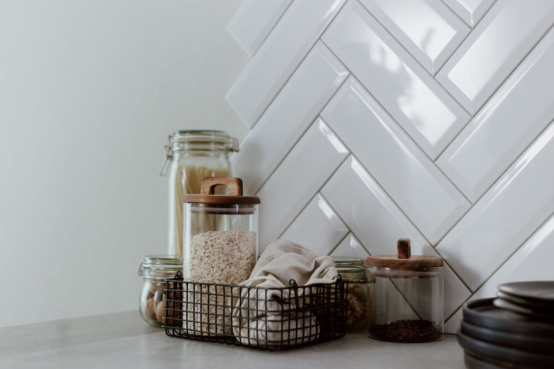 Glass storage jars with wooden lids holding oats, pasta and coffee beans beside a white tiled wall