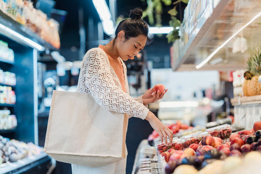 Woman in a white crochet cardigan holding a peach in a grocery store aisle with a large cream canvas tote bag over her shoulder