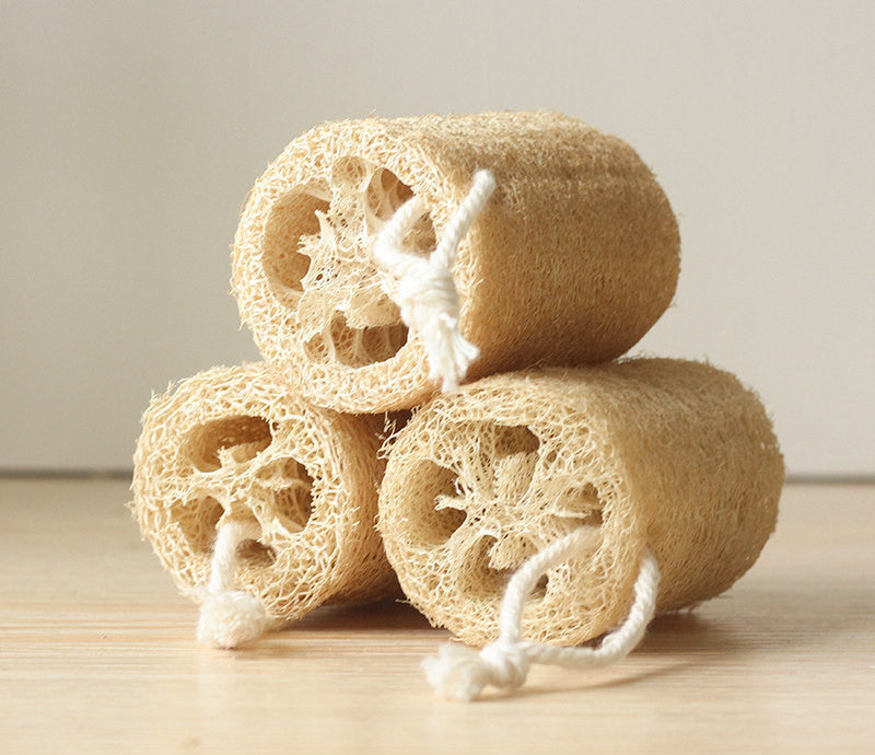 Three natural loofah scrubbers with cotton cords stacked on a light wooden surface
