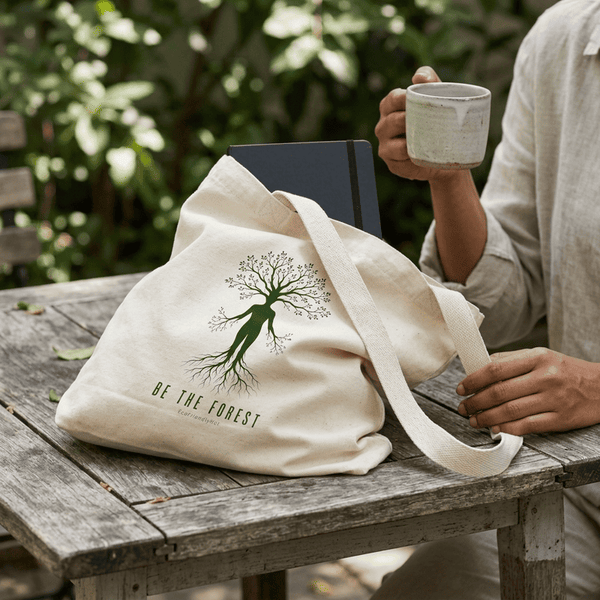 Cream canvas tote with a tree-shaped figure and the words Be the Forest, sitting on a wooden table with a notebook in it while a person holds a mug